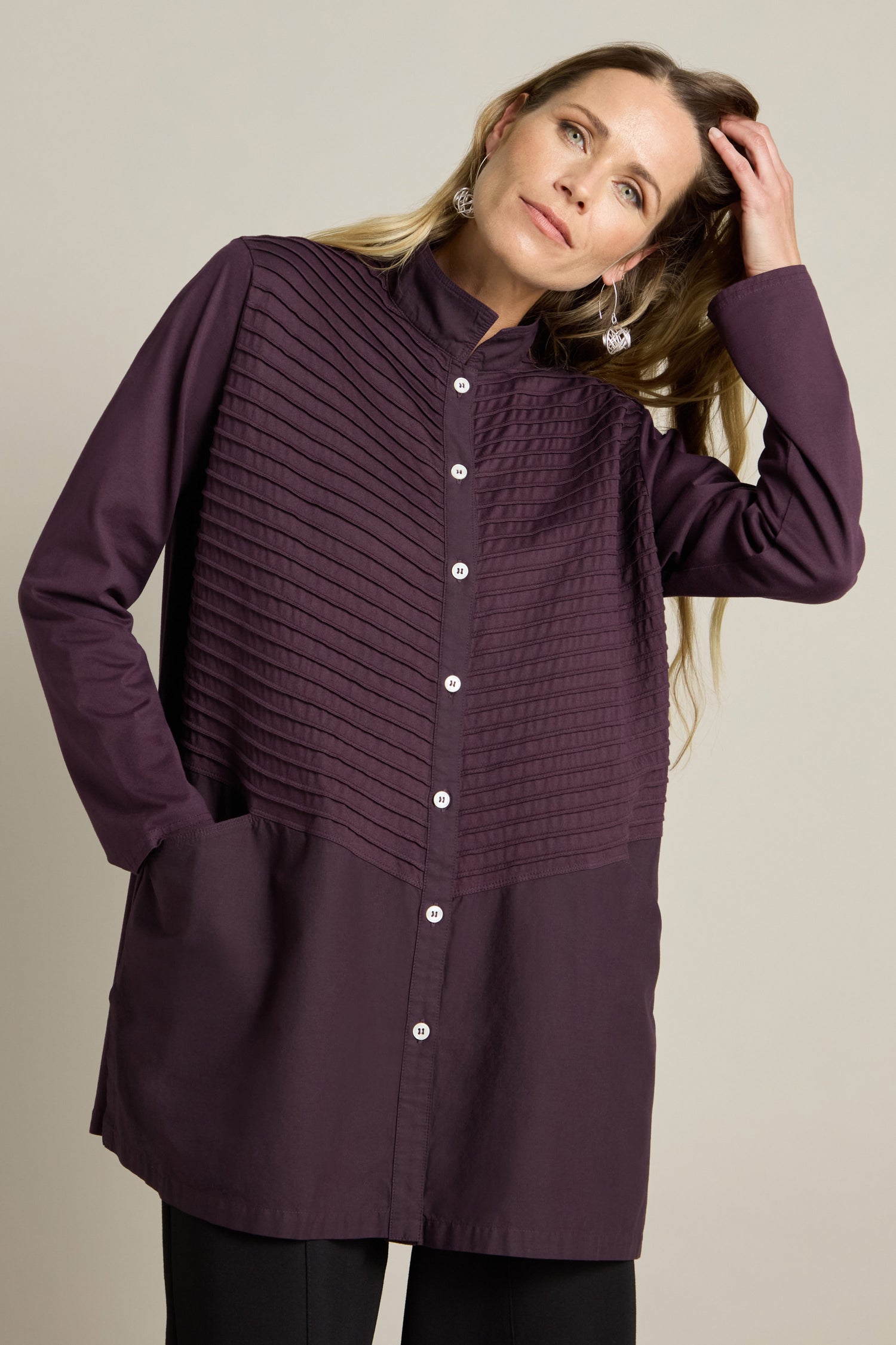 A woman wearing the Cotton Jersey And Calico Overshirt, with long sleeves and a dark purple hue, featuring a button-up design and embroidered placket along with textured detailing on the upper half, poses with her hands in her pockets and one hand touching her hair.