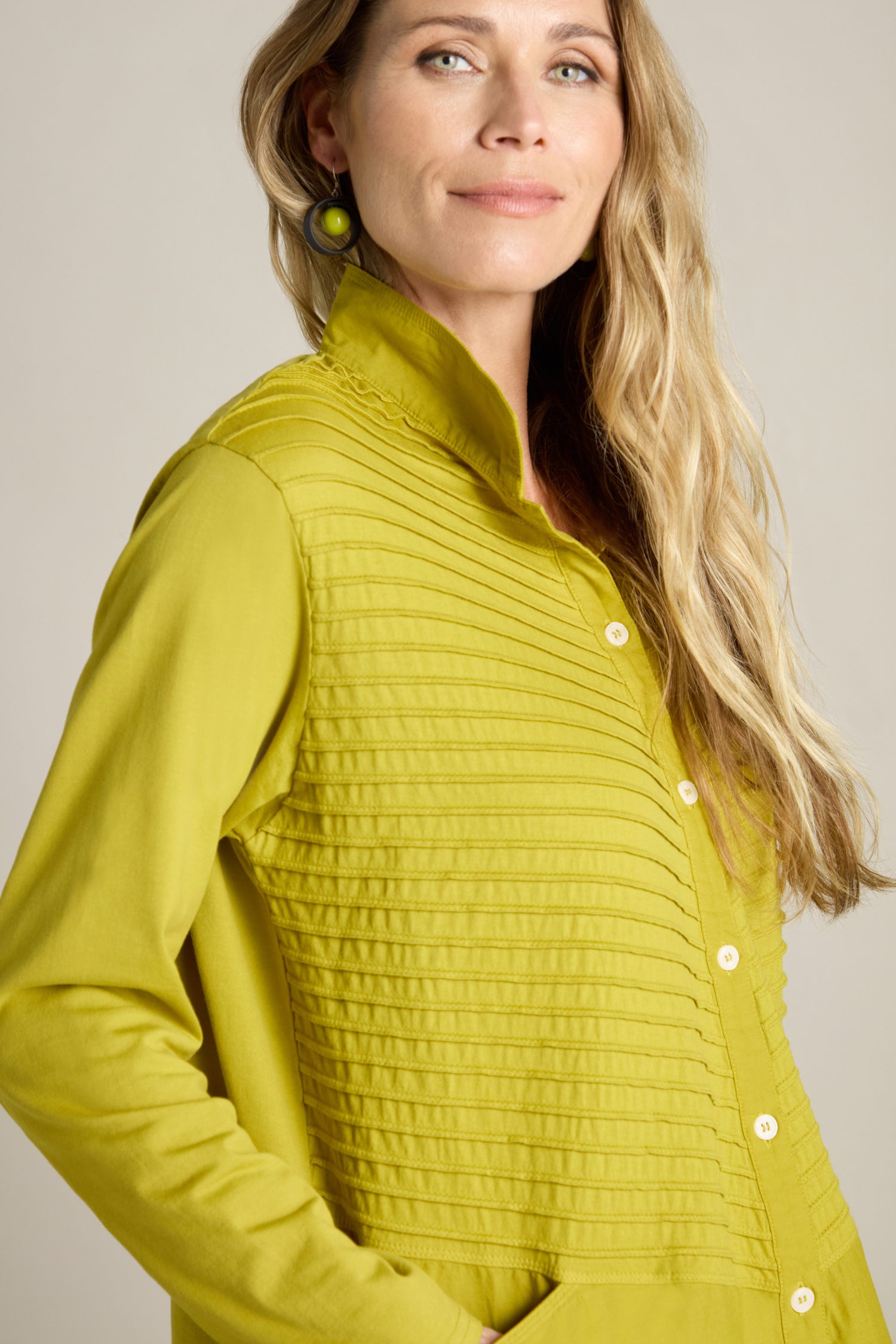 A person with long blonde hair wearing the Cotton Jersey And Calico Overshirt, featuring a green button-up design with vertical pleats on the front and an embroidered placket, looks at the camera.