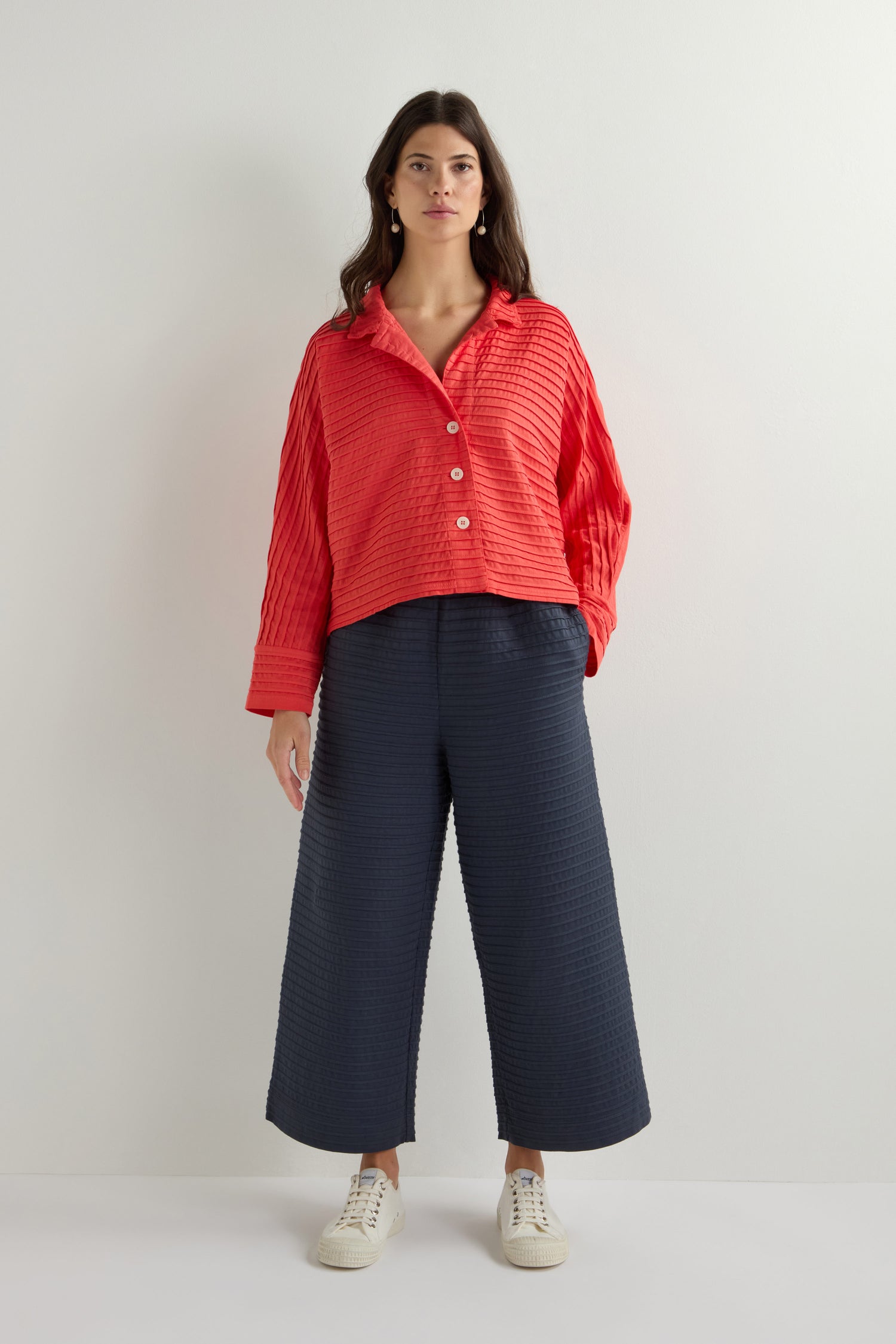 A person stands against a plain background, modeling the Yacco Maricard Jersey Pleat Short Boxy Jacket in red, paired with black wide-leg pants and white sneakers.