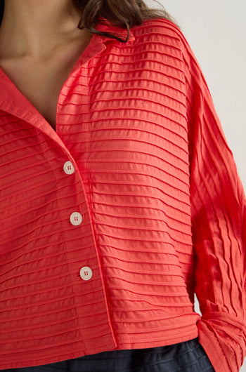 A person is wearing a red textured Jersey Pleat Short Boxy Jacket by Yacco Maricard, characterized by horizontal ribbing and white buttons. Only the upper torso is visible.