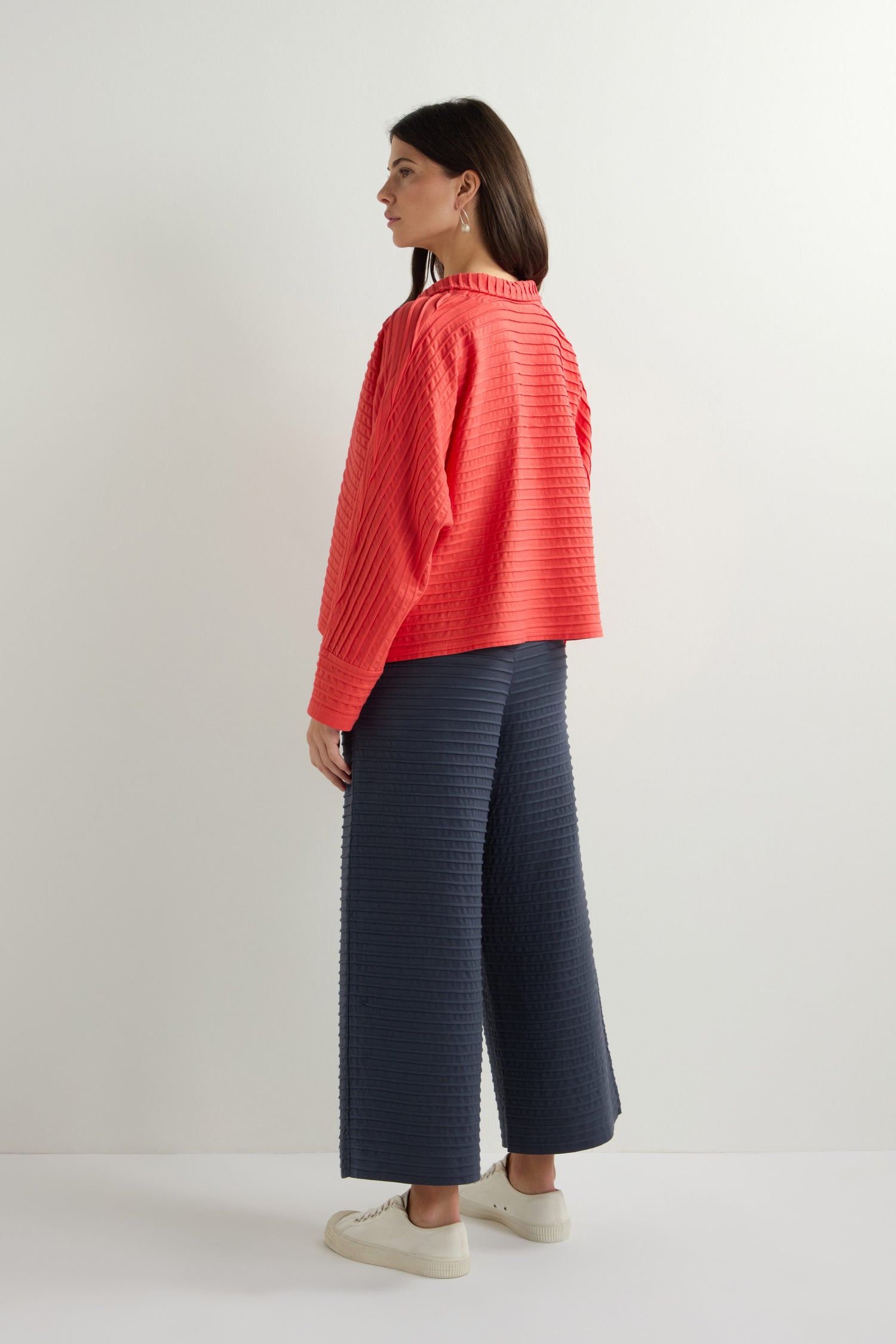 A woman in a coral-textured top and navy wide-leg pants stands against a plain white background, her gaze cast sideways, exuding the effortless elegance typical of Yacco Maricard's Jersey Pleat Short Boxy Jacket signature style.