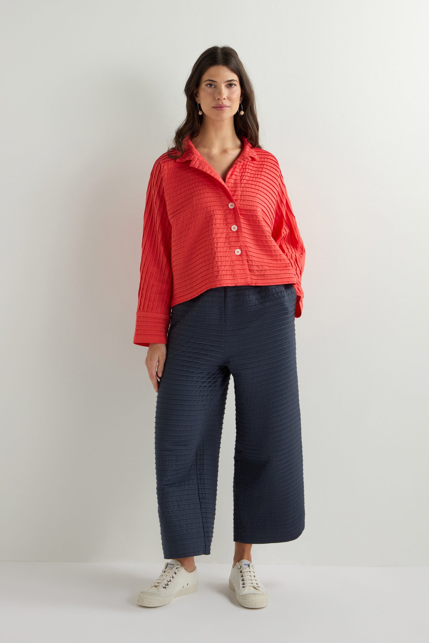 A woman in a red button-up shirt, navy wide-leg pants, and white sneakers stands against a plain light backdrop, effortlessly layering her look with a Jersey Pleat Short Boxy Jacket from Yacco Maricard.