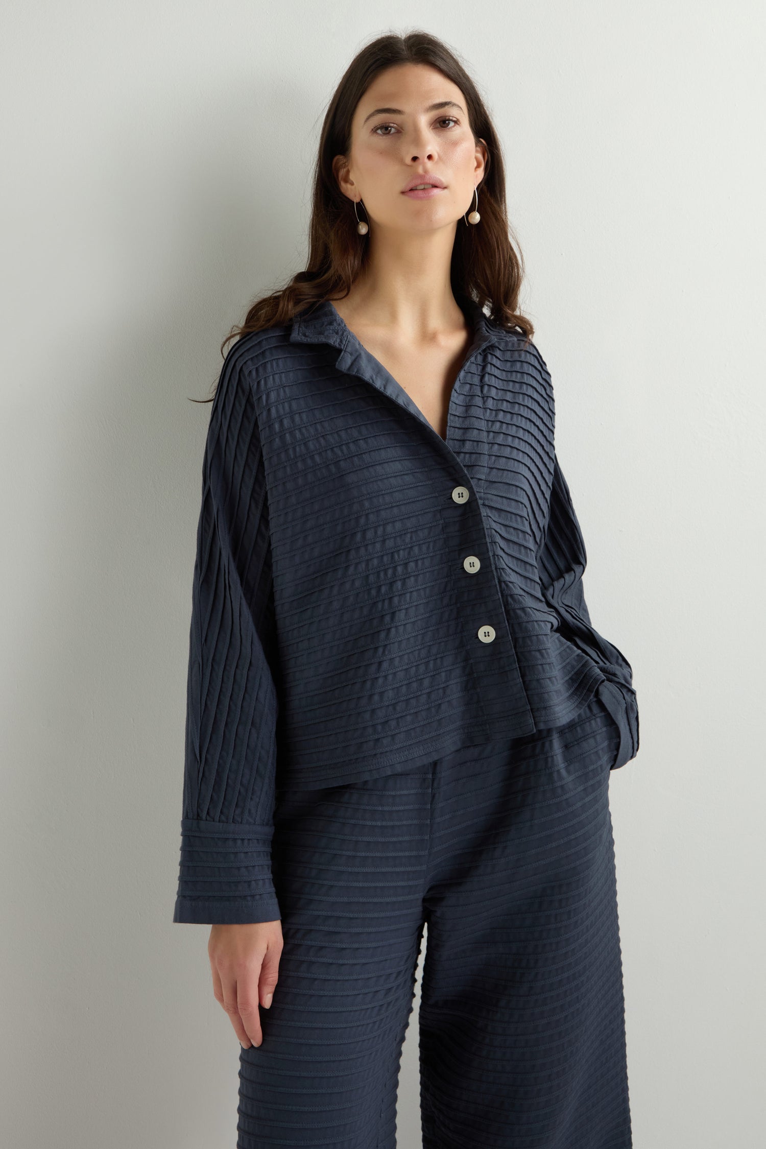 A person stands against a plain background, wearing a navy textured Jersey Pleat Short Boxy Jacket from Yacco Maricard with matching wide-leg pants, hands in pockets.