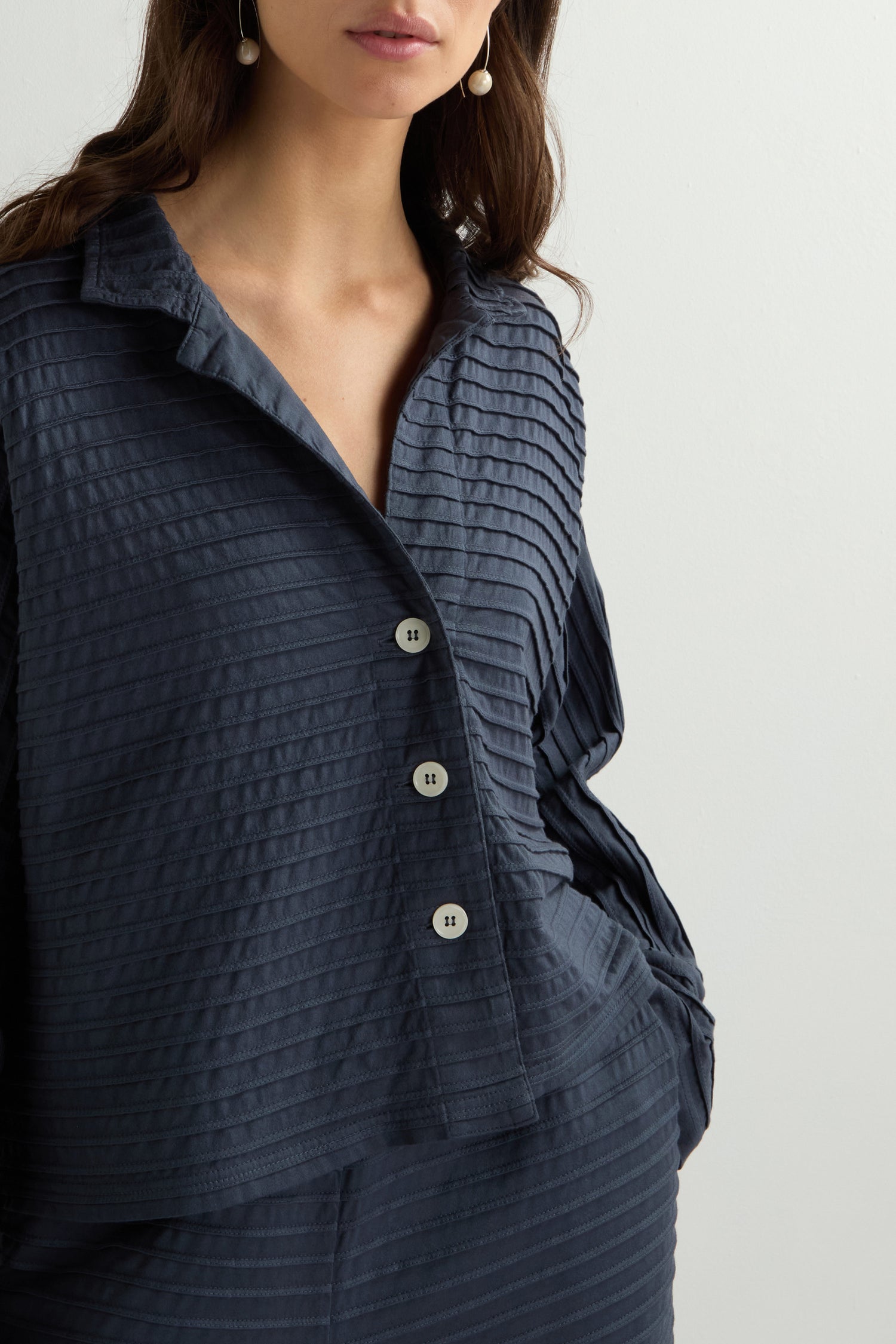 A woman stands elegantly against a white backdrop, wearing a textured dark gray button-up blouse with matching pants. Her long brown hair complements the outfit, subtly reflecting the chic style of a Jersey Pleat Short Boxy Jacket.