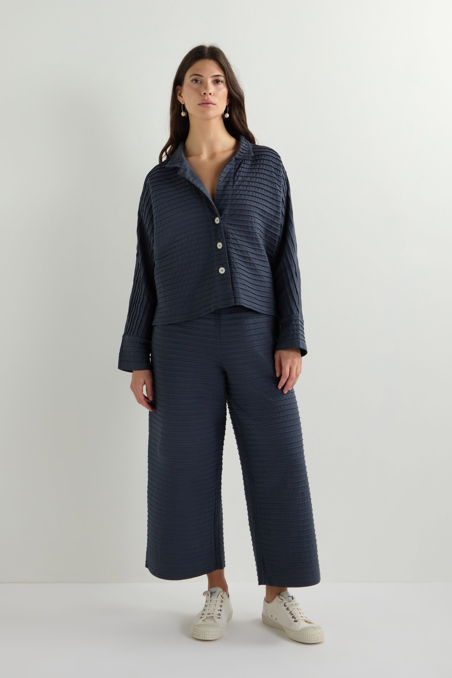 A woman poses against a plain backdrop in a textured dark blue outfit, inspired by Yacco Maricard, featuring the Jersey Pleat Short Boxy Jacket and matching wide-leg pants. White sneakers complete her look.