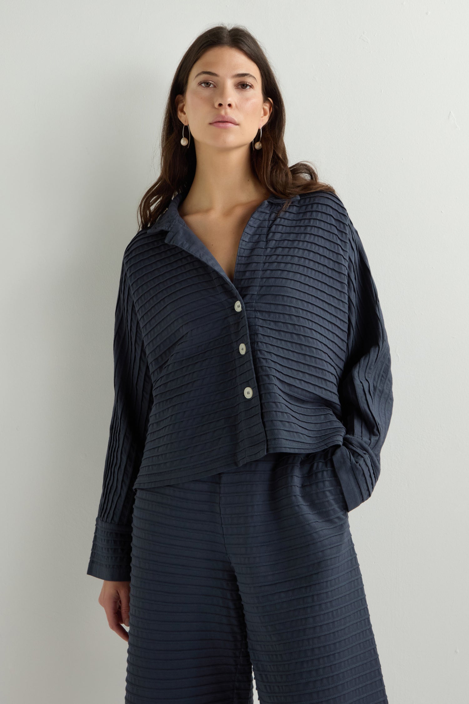 A woman wears a dark, textured Jersey Pleat Short Boxy Jacket and matching pants, standing against a plain background.