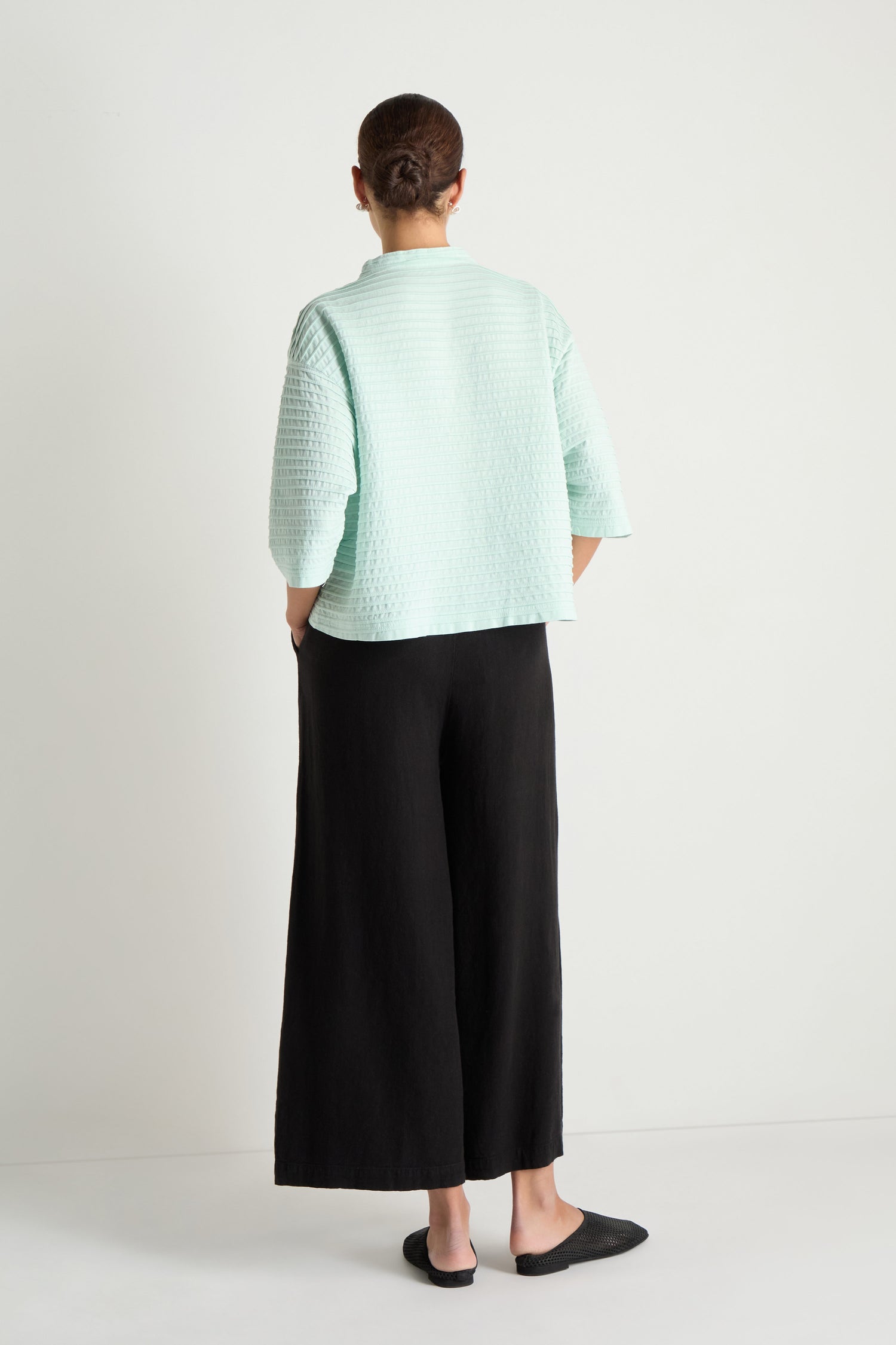 A person stands facing away, wearing the Funnel Neck Stone Blue Cotton Jacket with three-quarter sleeves, wide black pants, and black slip-on shoes. The jacket's boxy silhouette contrasts against the plain white background.