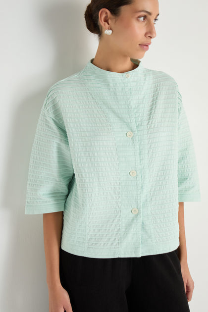 A woman wearing a Funnel Neck Stone Blue Cotton Jacket with wide sleeves and a boxy silhouette, paired with black pants, stands against a white wall and looks to the side.