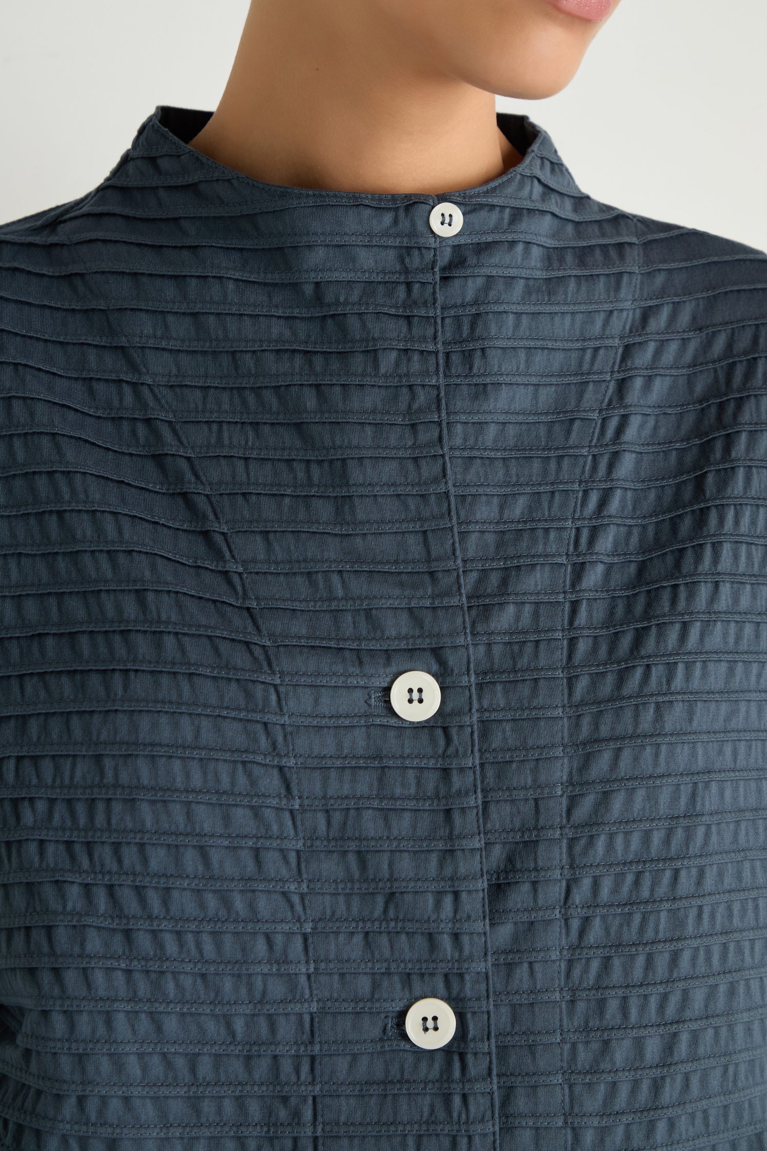 Close-up of a person wearing the Funnel Neck Stone Blue Cotton Jacket featuring a dark textured fabric, stand collar, white buttons, and boxy silhouette; the person’s face is partially out of frame.