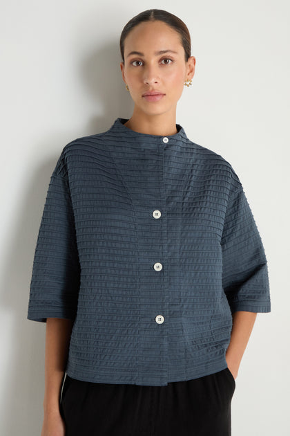 A woman stands against a plain white wall wearing the Funnel Neck Stone Blue Cotton Jacket, featuring a boxy silhouette and elbow-length sleeves.