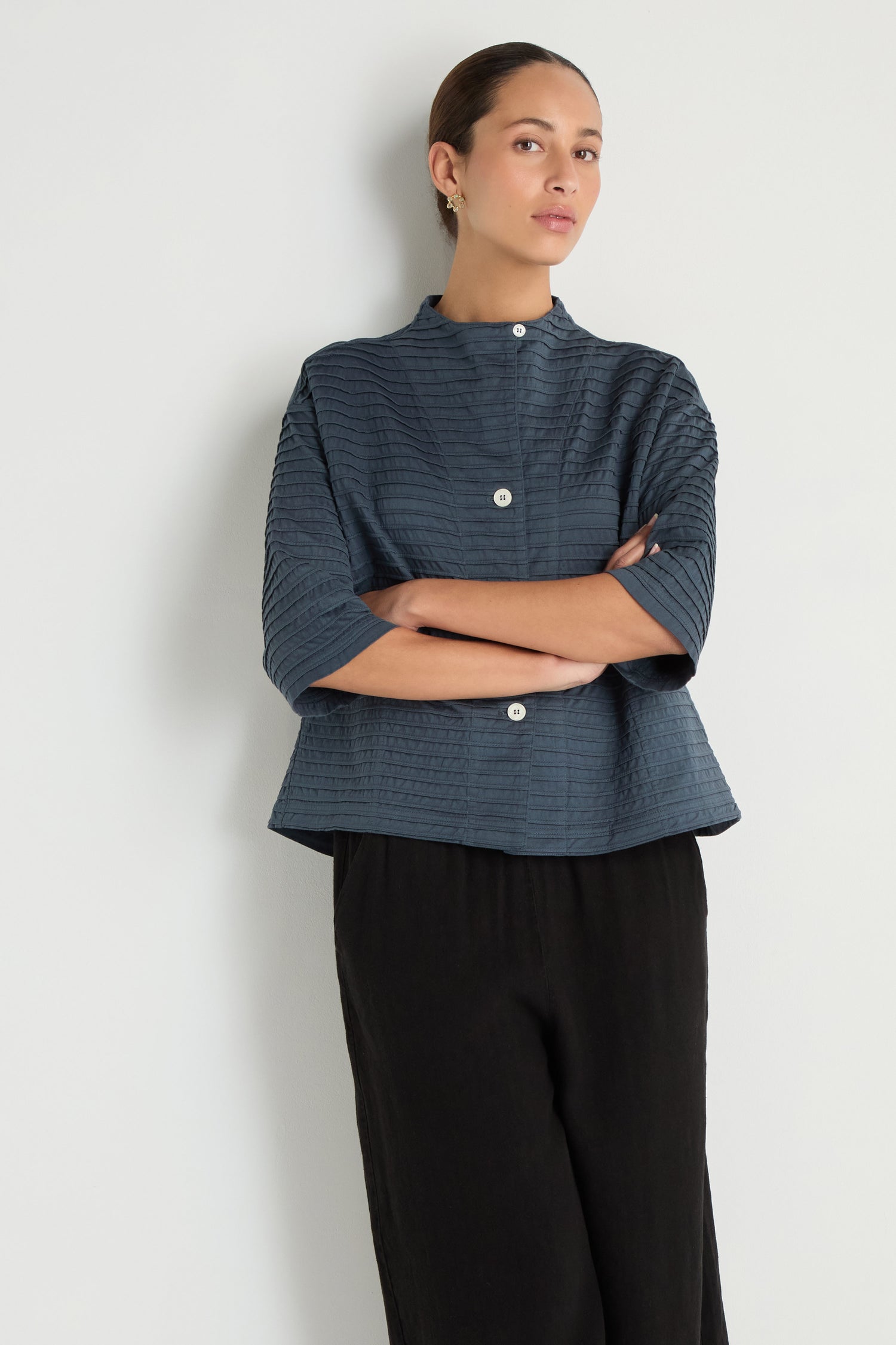 A person stands against a white wall, arms crossed, wearing a Funnel Neck Stone Blue Cotton Jacket with a boxy silhouette and three-quarter sleeves, paired with loose black pants.