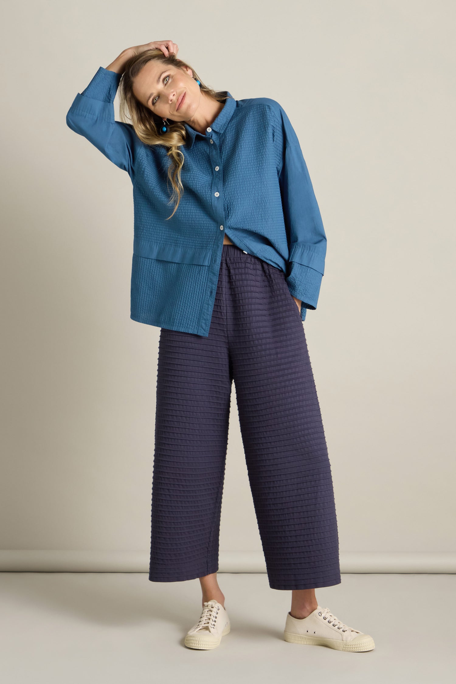 A woman in a blue button-up shirt and navy Cotton Jersey Pleat Trouser, echoing a loungewear-inspired vibe, poses with one hand resting on her head. She stands against a plain light-colored background and complements her outfit with white sneakers.