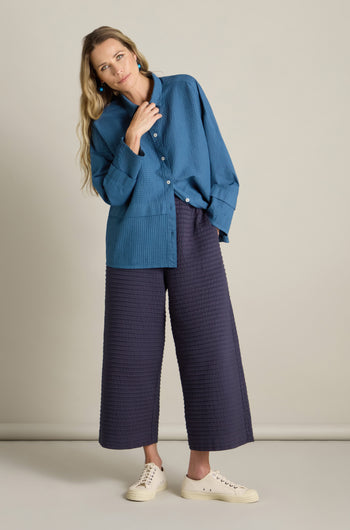 A woman wearing a loose blue button-up shirt and wide-legged, loungewear-inspired Cotton Jersey Pleat Trousers from Yacco Maricard stands against a plain background. She has long hair and is sporting white sneakers.