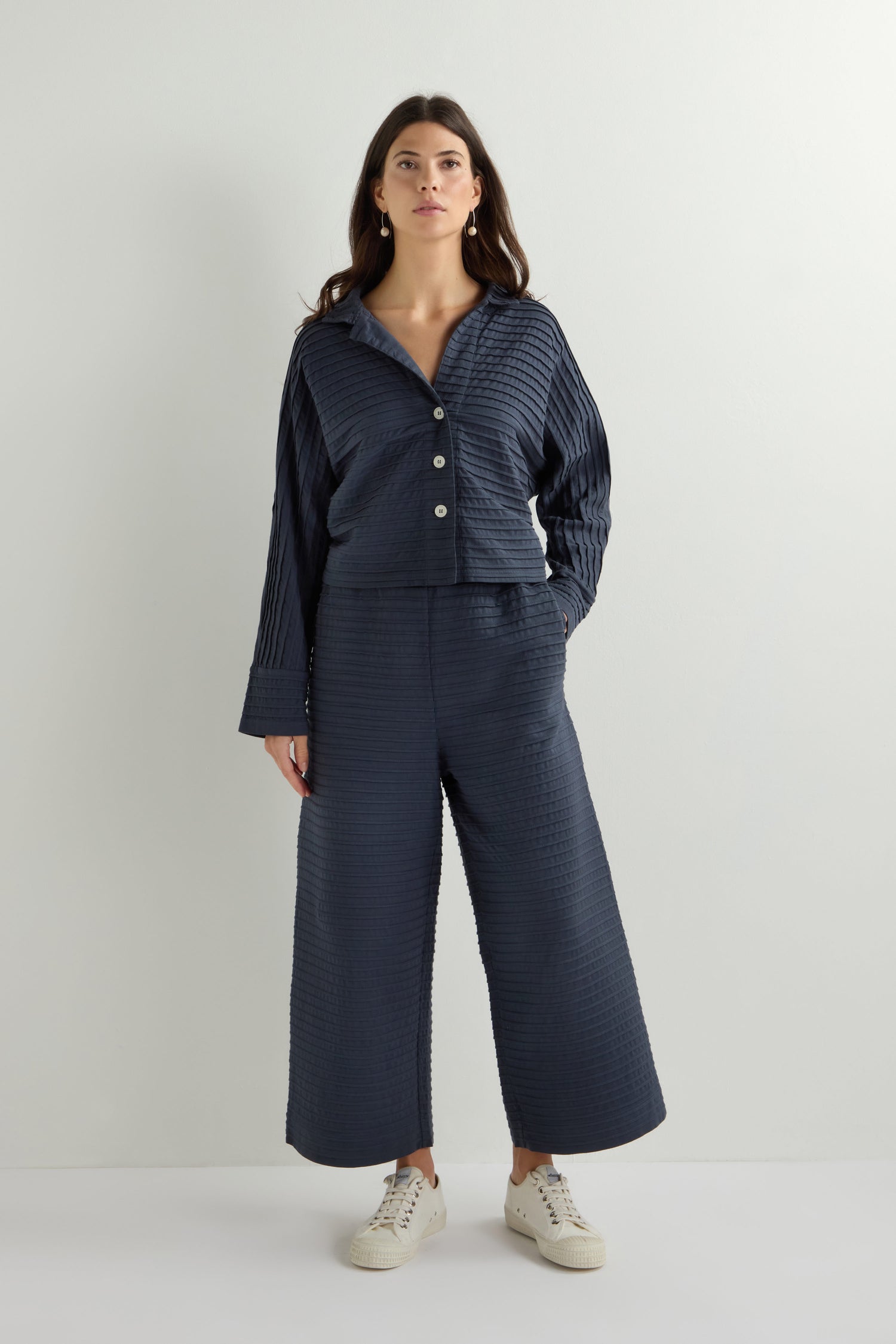 A woman in navy, loungewear-inspired styling featuring the Pleated Jersey Trouser and a button-up top stands against a white background, paired with white sneakers.