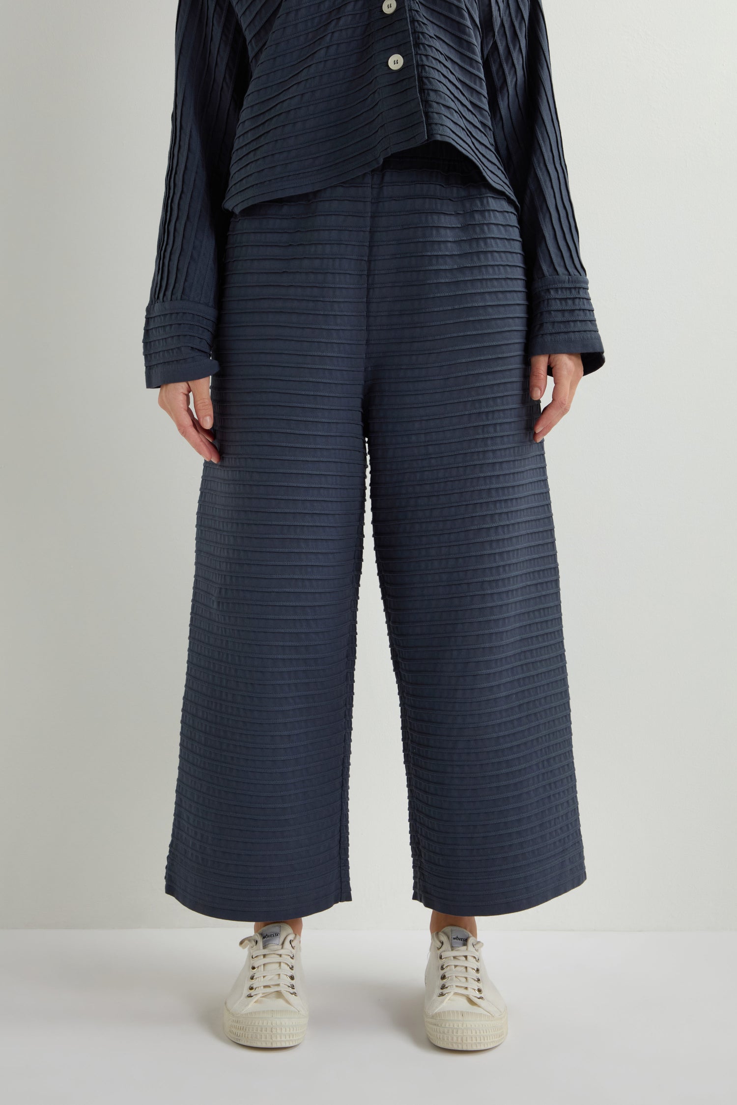 Against a plain backdrop, someone wears a textured navy ensemble with Pleated Jersey Trousers and white sneakers, showcasing loungewear-inspired style through its wide-legged design.