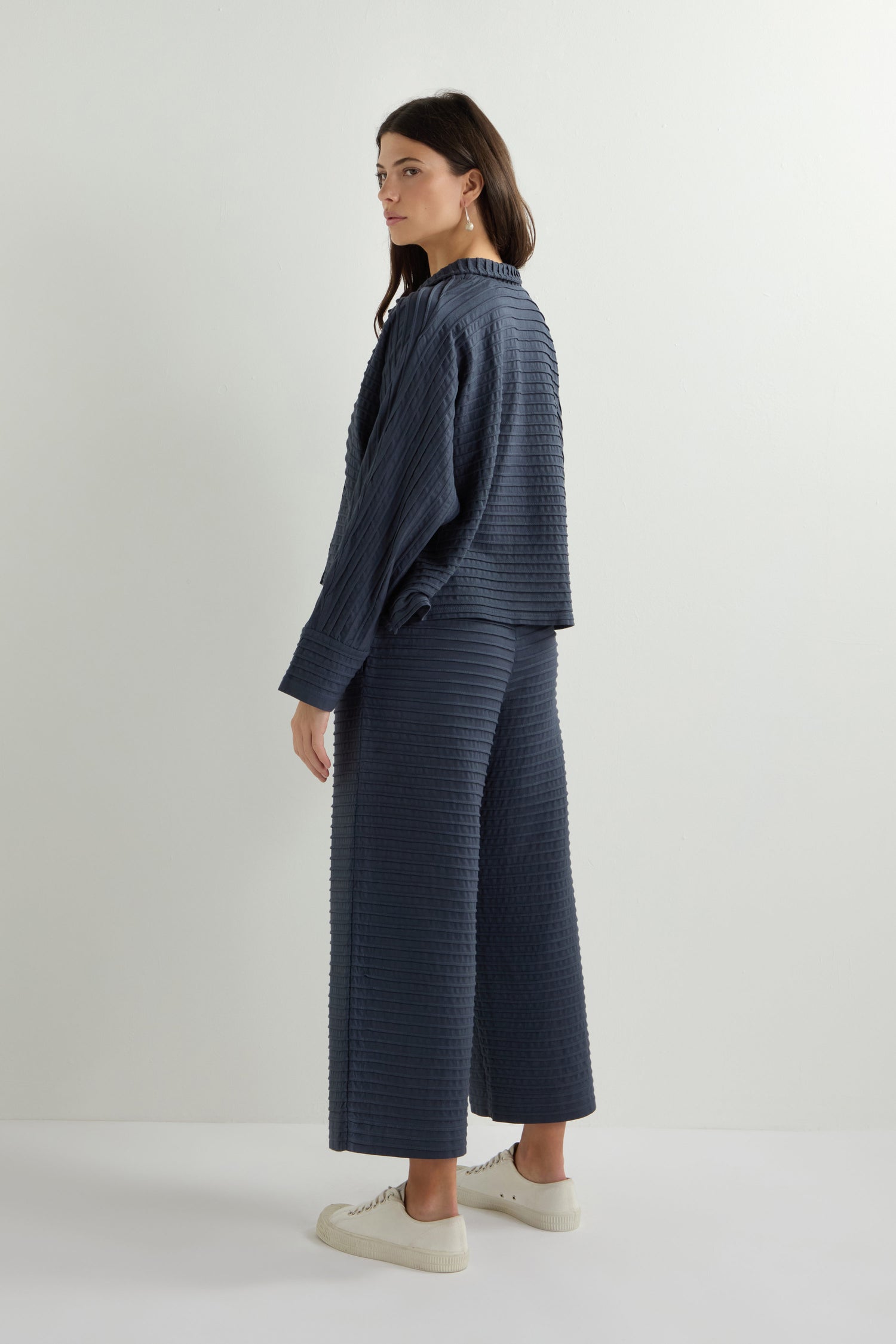 A person stands sideways wearing a textured blue Pleated Jersey Trouser with a matching jacket and white sneakers, creating loungewear-inspired styling that effortlessly blends comfort with chic appeal.