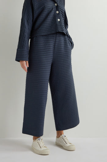 A person dons a navy textured long-sleeve shirt paired with Pleated Jersey Trousers and white sneakers against a plain backdrop, showcasing a chic, relaxed loungewear-inspired style.