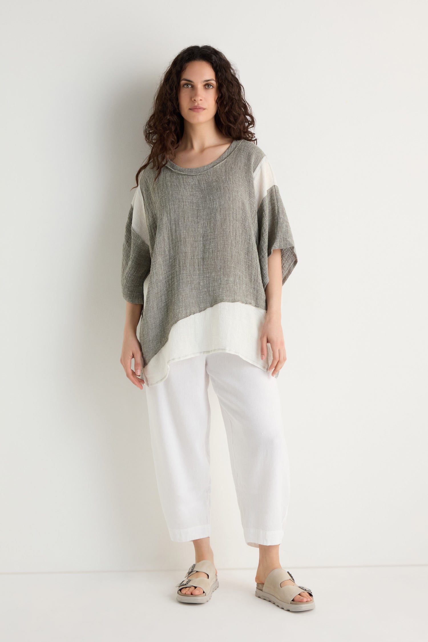 A woman stands against a plain white background wearing the Linen Blend Boxy Tunic with contrast panel detailing, paired with white pants and beige sandals.