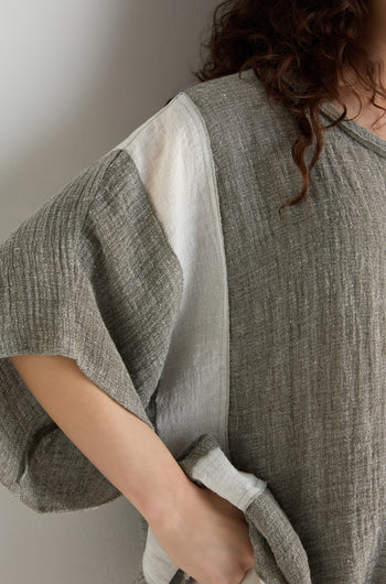 Wearing a Linen Blend Boxy Tunic with gray and white contrast panels, this person stands against a light-colored wall. Only the upper body is partially visible.