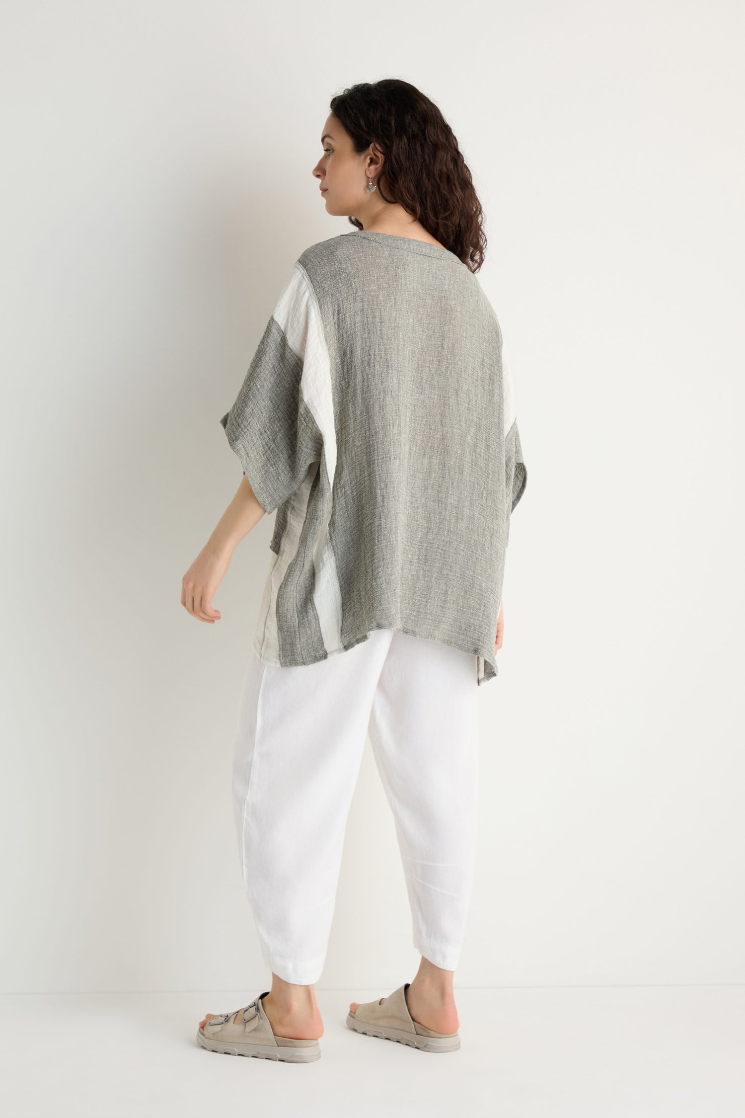 A person with curly dark hair stands facing a white wall, wearing the Linen Blend Boxy Tunic in gray and white with contrast panels, paired with white pants and beige sandals.
