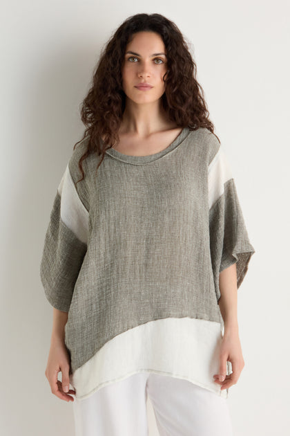 Woman with long curly hair wearing the Linen Blend Boxy Tunic—loose-fitting, short-sleeve, gray and white with contrast panel detailing—paired with white pants, standing against a plain light background.