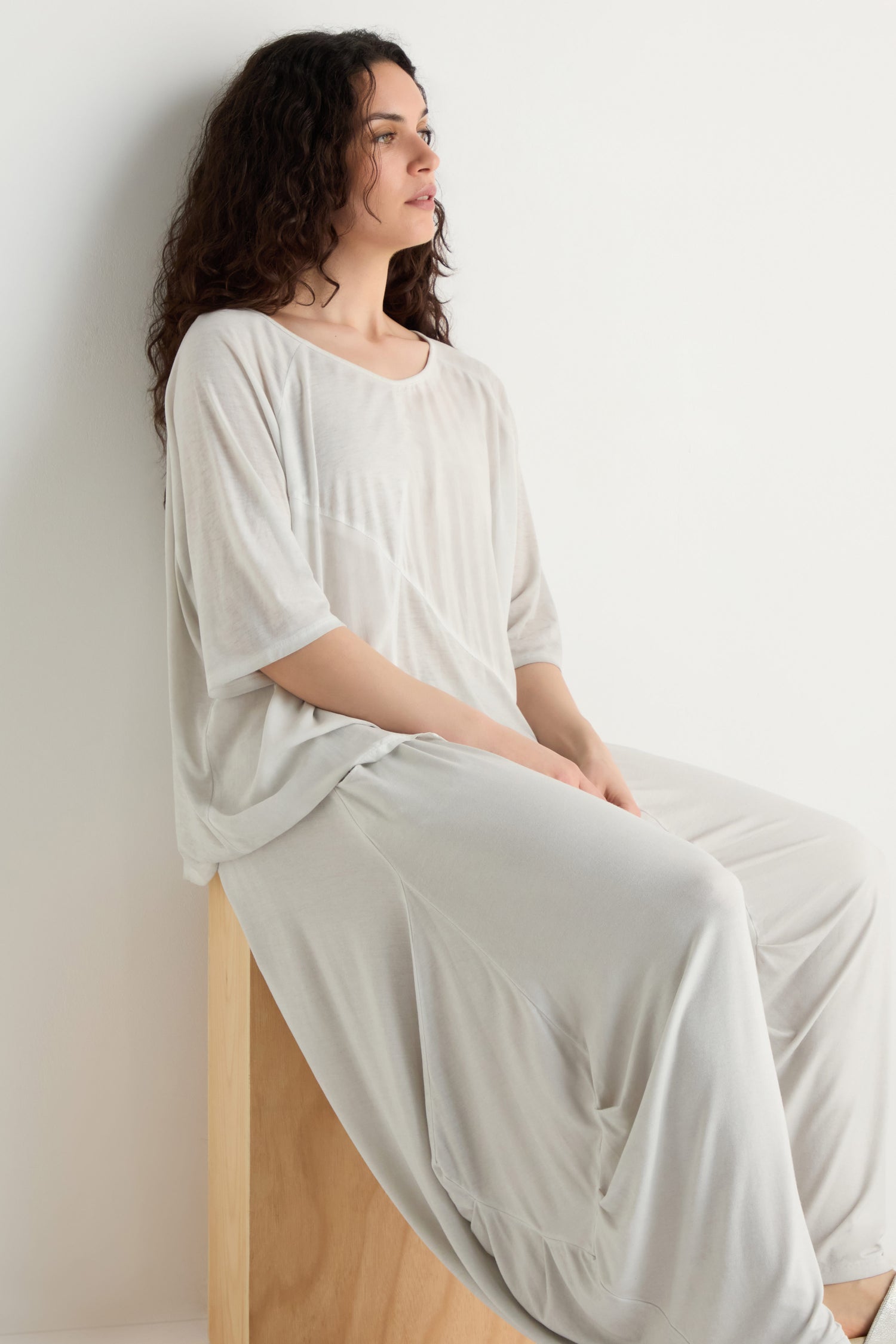 A person with long curly hair sits on a wooden stool, wearing a loose light beige top and Soft Drape Jersey Trousers with a relaxed wide-leg fit, set against a plain white background.