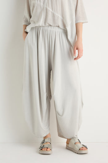 A person stands against a plain white background wearing loose, light gray Soft Drape Jersey Trousers, a matching top, and gray sandals.