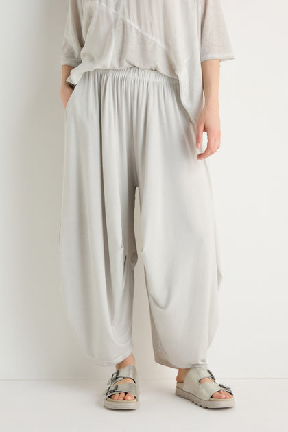 A person stands against a plain white background wearing loose, light gray Soft Drape Jersey Trousers, a matching top, and gray sandals.