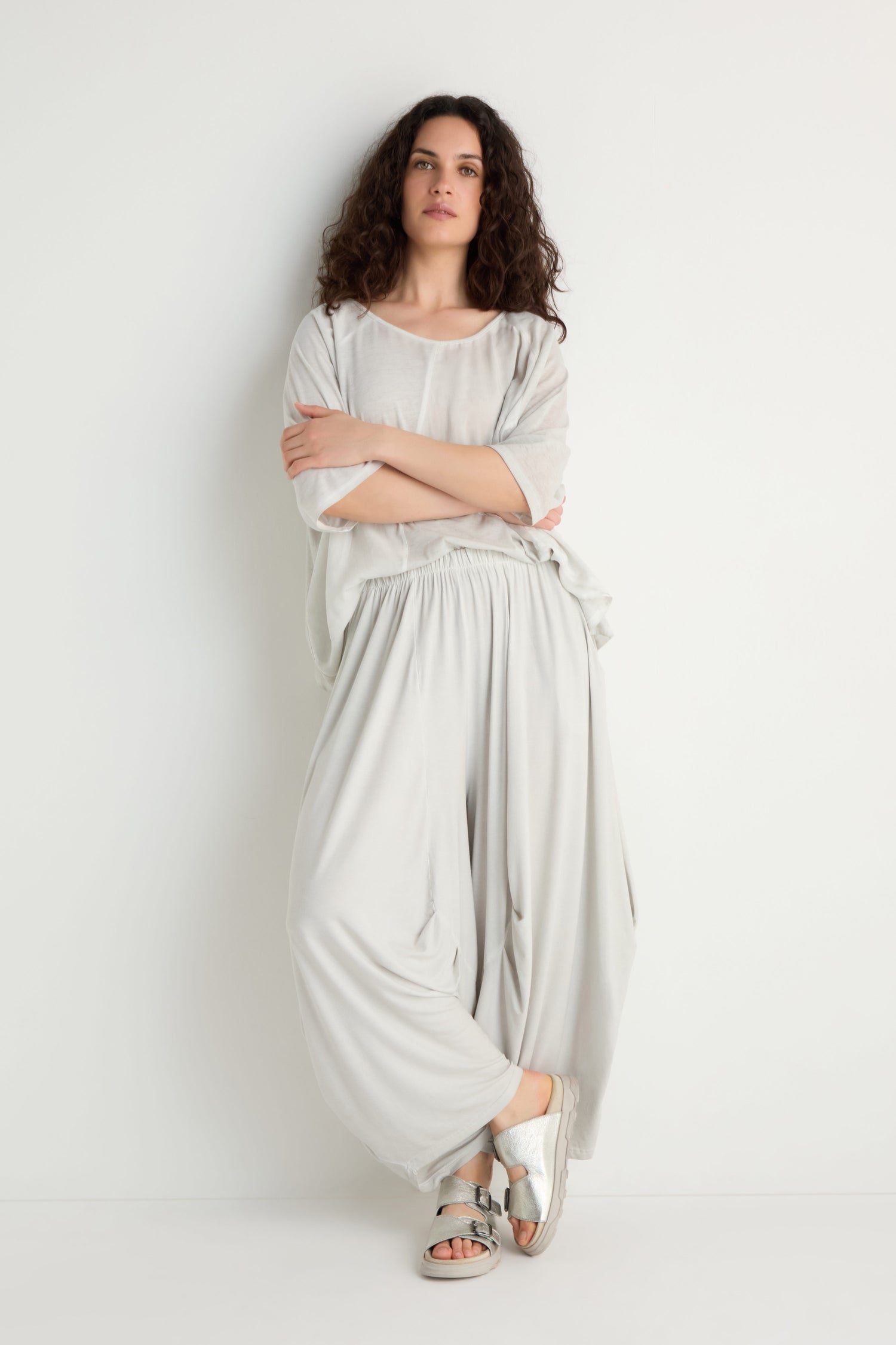 A person with curly hair stands against a white wall, wearing relaxed fit Soft Drape Jersey Trousers in light grey with a matching top and silver sandals, arms crossed and one leg slightly bent.