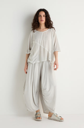 A woman stands against a white wall, wearing the Seam Detail Jersey Top—a loose viscose jersey with asymmetric seam details—paired with matching draped pants and gray sandals for a relaxed silhouette.