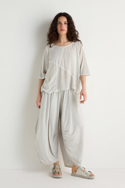A woman stands against a white wall, wearing the Seam Detail Jersey Top—a loose viscose jersey with asymmetric seam details—paired with matching draped pants and gray sandals for a relaxed silhouette.