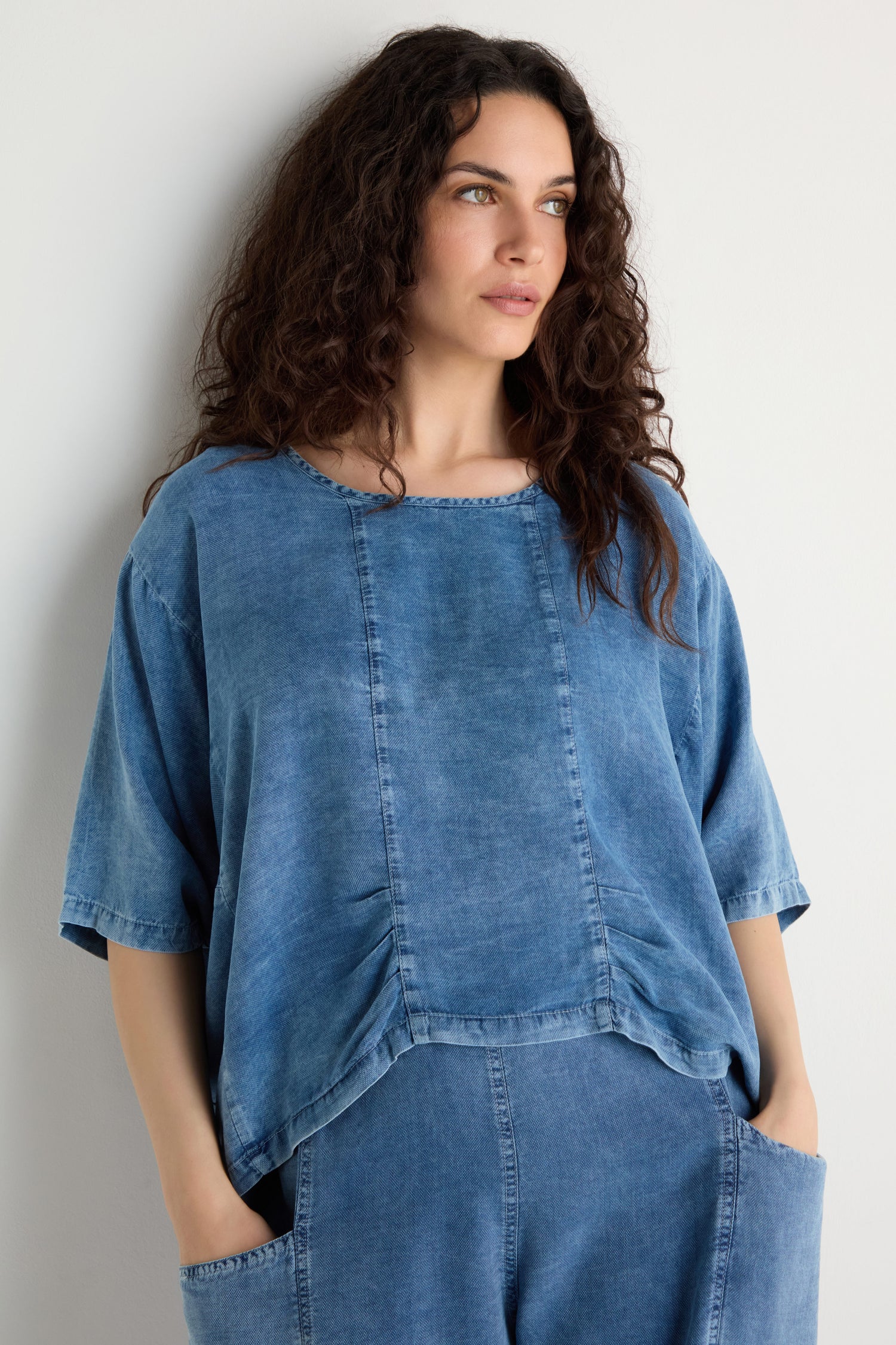 A woman with long curly hair, hands in her pockets, stands against a plain white wall wearing the Lyocell Panelled Boxy Top and matching pants.