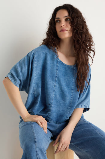 A woman with long curly hair sits on a wooden stool against a plain light background, wearing the Lyocell Panelled Boxy Top and matching denim pants with panelled details.