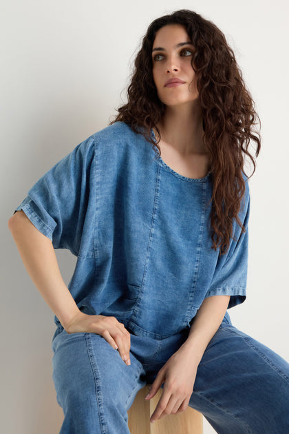 A woman with long curly hair sits on a wooden stool against a plain light background, wearing the Lyocell Panelled Boxy Top and matching denim pants with panelled details.