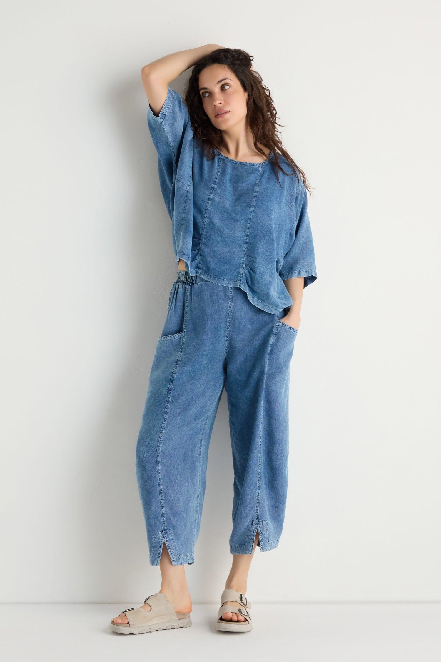 A woman poses against a white wall, wearing Lyocell Slit Hem Trousers in blue with a relaxed fit and slit hem, paired with a loose denim top and beige sandals, resting one hand on her head.