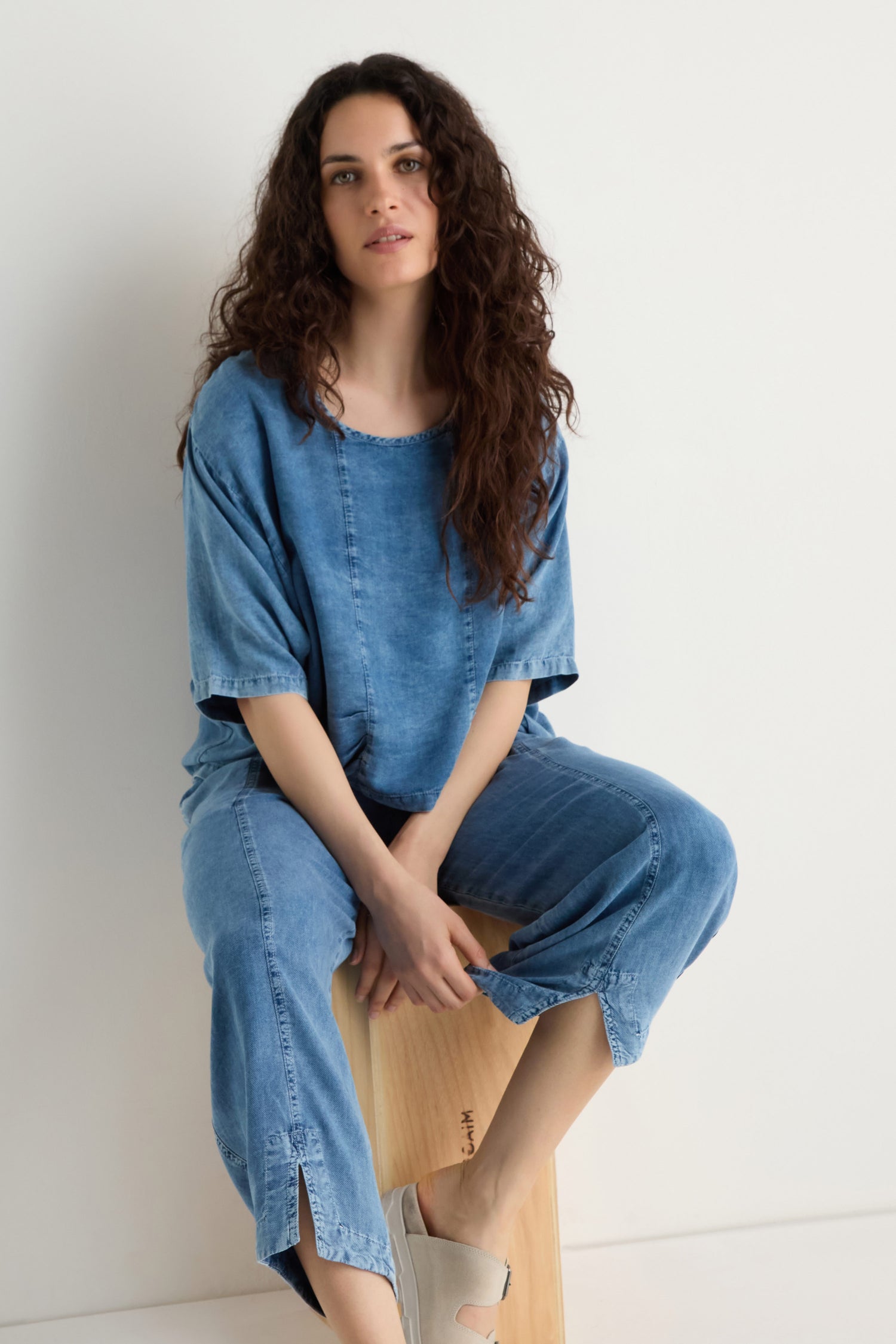 A woman with long curly hair wears a loose denim top and Lyocell Slit Hem Trousers with a relaxed leg, seated on a wooden stool against a plain white background.