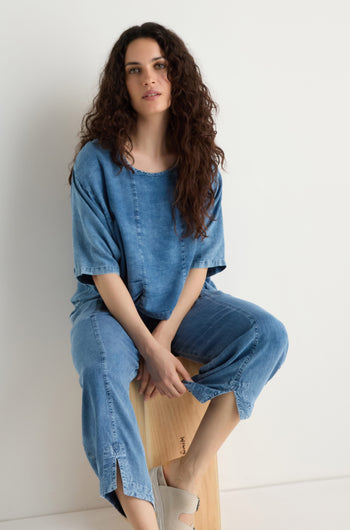 A woman with long curly hair wears a loose denim top and Lyocell Slit Hem Trousers with a relaxed leg, seated on a wooden stool against a plain white background.