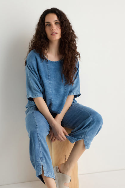 A woman with long curly hair wears a loose denim top and Lyocell Slit Hem Trousers with a relaxed leg, seated on a wooden stool against a plain white background.