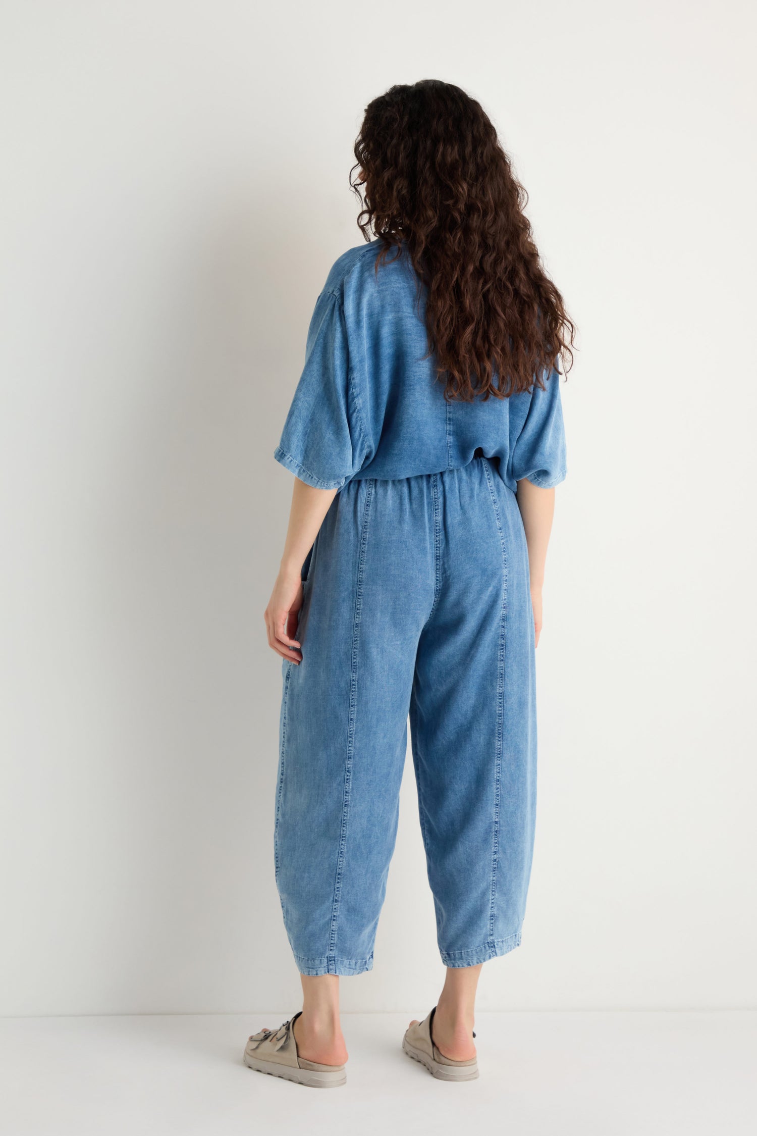 A person with long curly hair faces away, wearing Lyocell Slit Hem Trousers and beige slip-on shoes, paired with a loose-fitting blue denim jumpsuit against a plain white background.