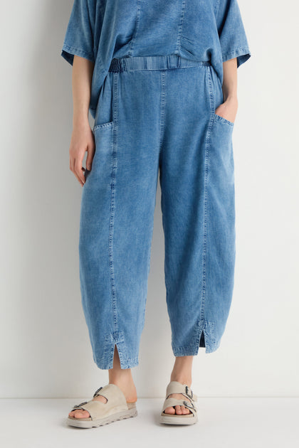 Model wearing Lyocell Slit Hem Trousers in a relaxed fit, with front seams and hands in pockets, paired with beige sandals and standing against a plain white background.