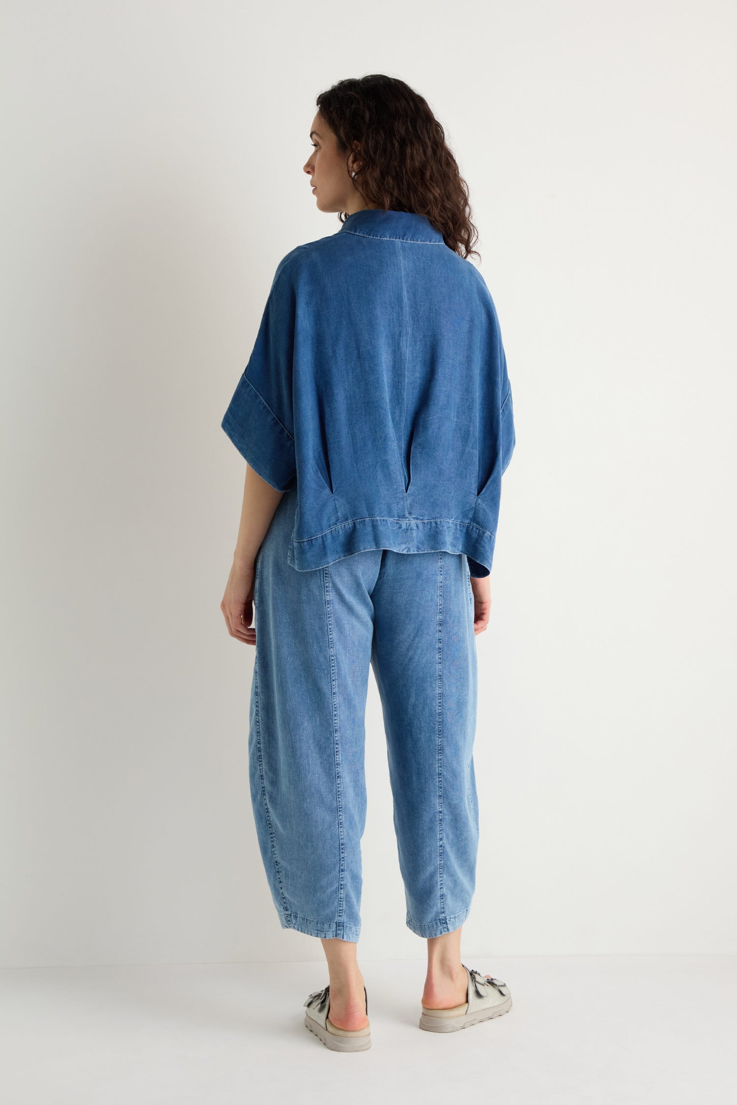 A woman faces away against a white background, wearing the Lyocell Boxy Shirt in blue with a relaxed fit, matching blue denim pants, and beige shoes.