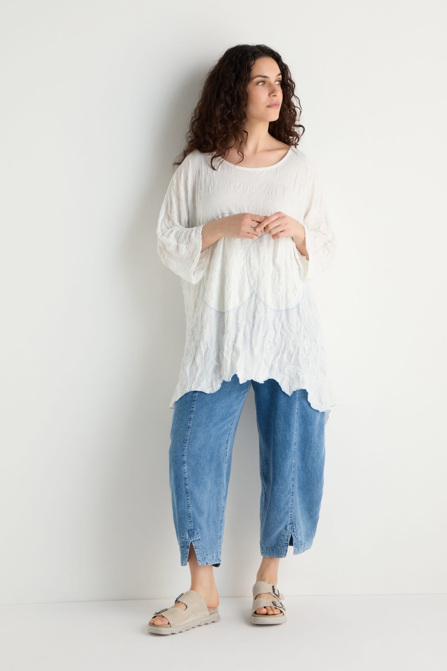 A person with curly hair stands against a white wall, wearing the Embroidered Circles Crinkle Tunic, blue cropped jeans, and beige sandals.