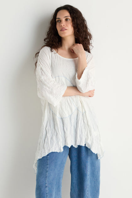 A woman with long curly hair stands against a white background, wearing the Embroidered Circles Crinkle Tunic in white and blue wide-leg jeans, looking to the side with one hand touching her face.
