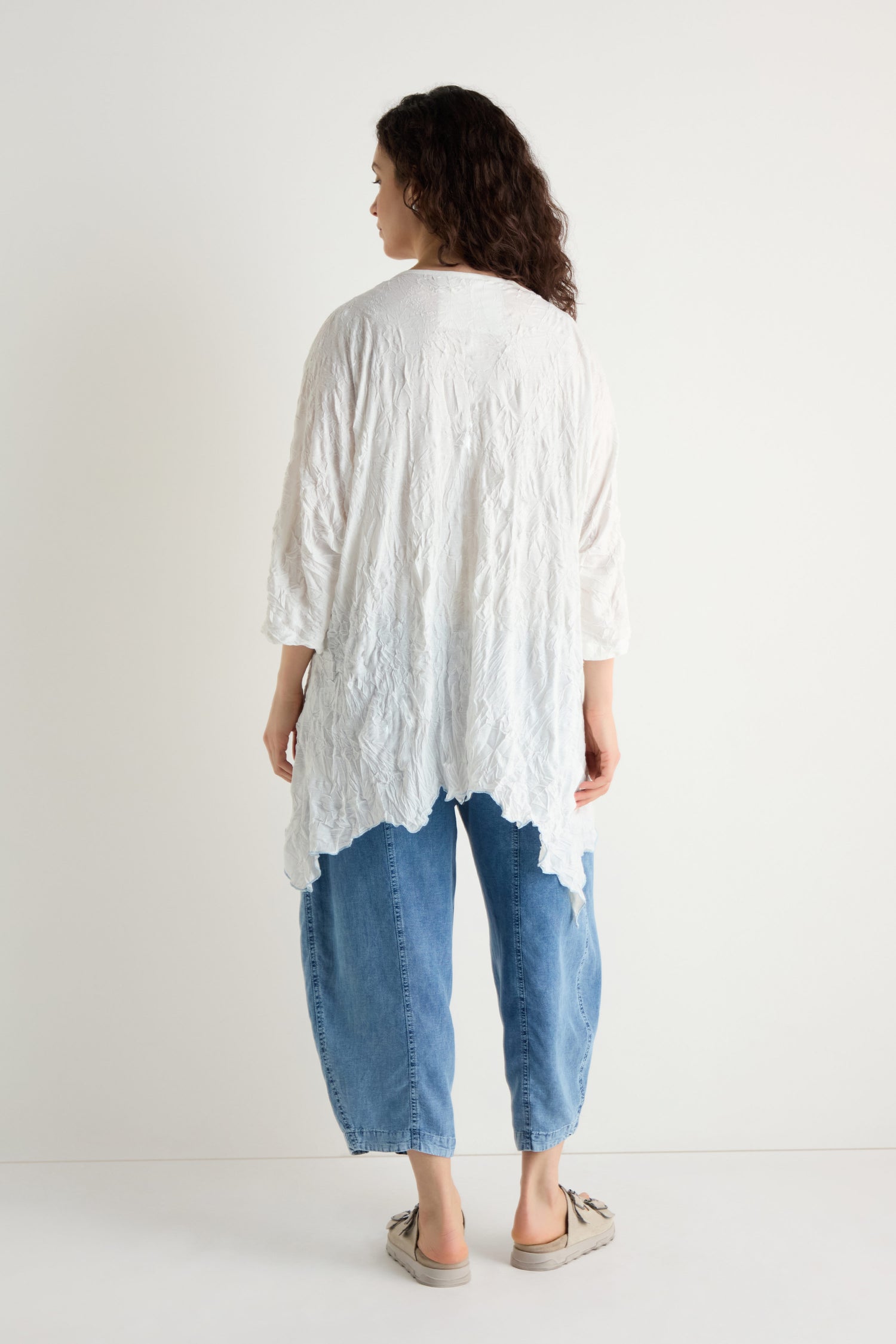 A person faces away, wearing the Embroidered Circles Crinkle Tunic in white, paired with light blue baggy jeans and beige slip-on shoes, set against a plain white background.