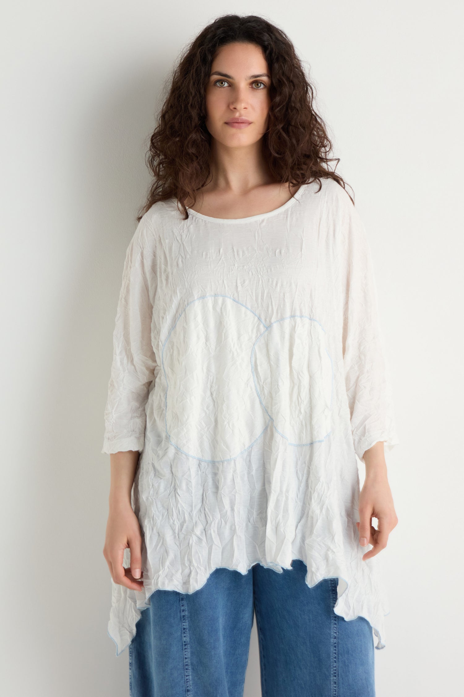 A woman with curly hair models the Embroidered Circles Crinkle Tunic, a loose white top with circle embroidery, paired with blue jeans against a plain white background.