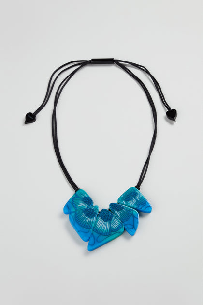 The Primula Resin Necklace by Zsiska, with three blue wing-shaped pendants on a black adjustable cord, is displayed against a light gray background—a striking piece of modern jewellery.