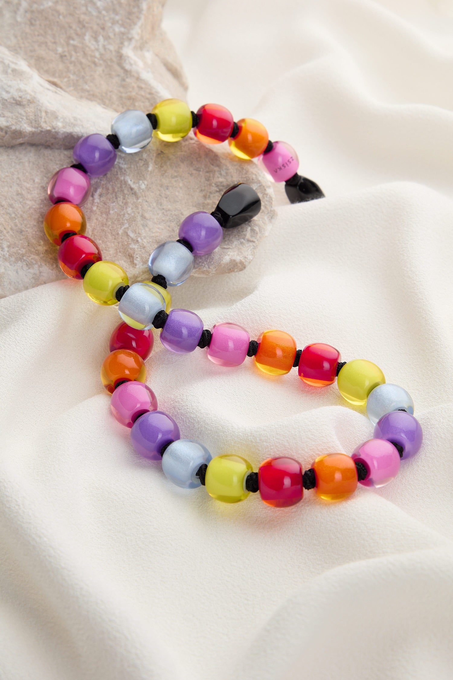 A Colourful Beads Necklace featuring vibrant resin beads in shades of pink, purple, yellow, orange, and blue, displayed on a white fabric background near some rocks. The Colourful Beads Necklace is elegantly designed with a convenient magnetic closure.