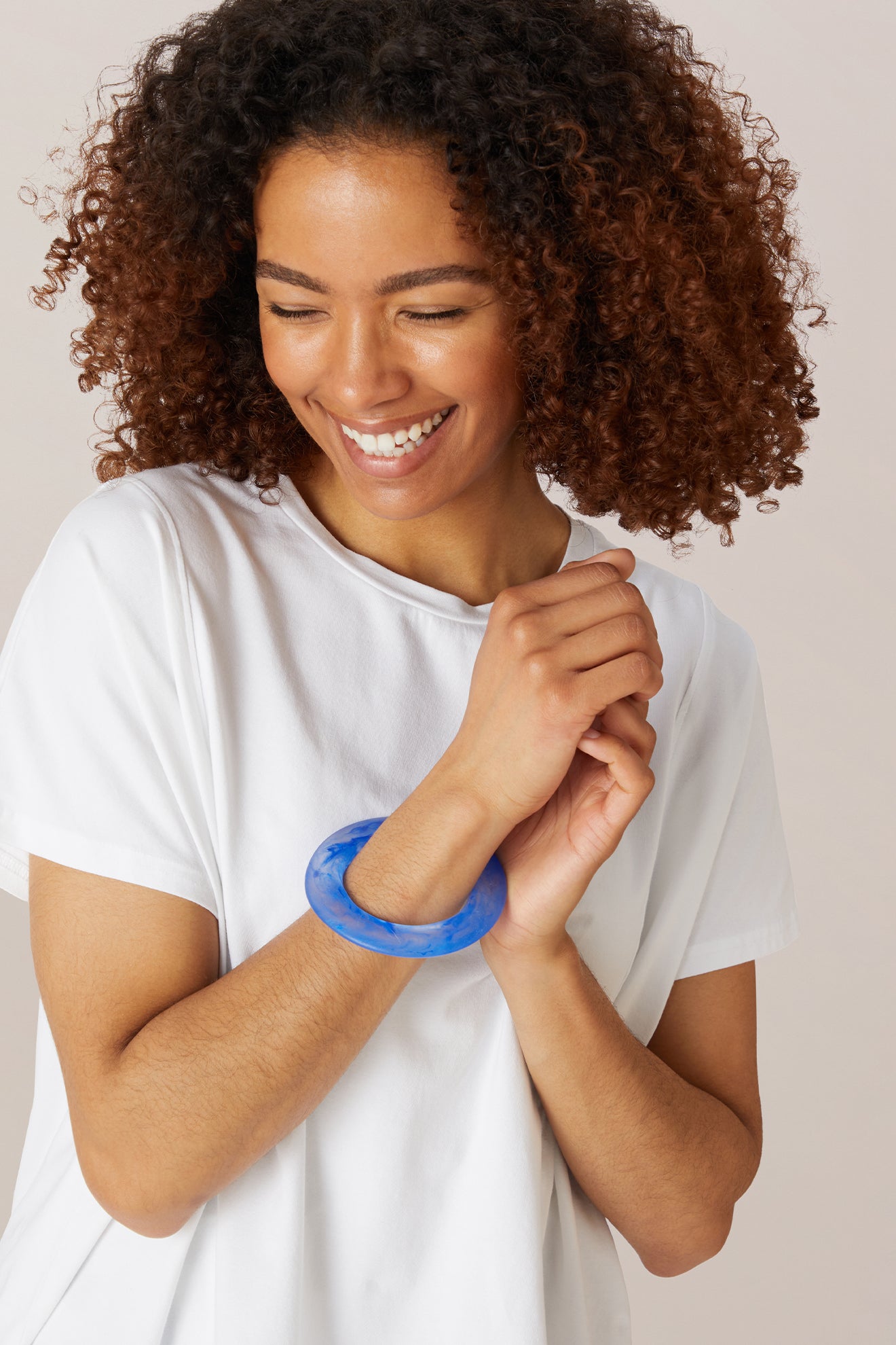 A woman smiles while looking down, hands clasped in front of her, wearing a white shirt and the Marbelled Bracelet.