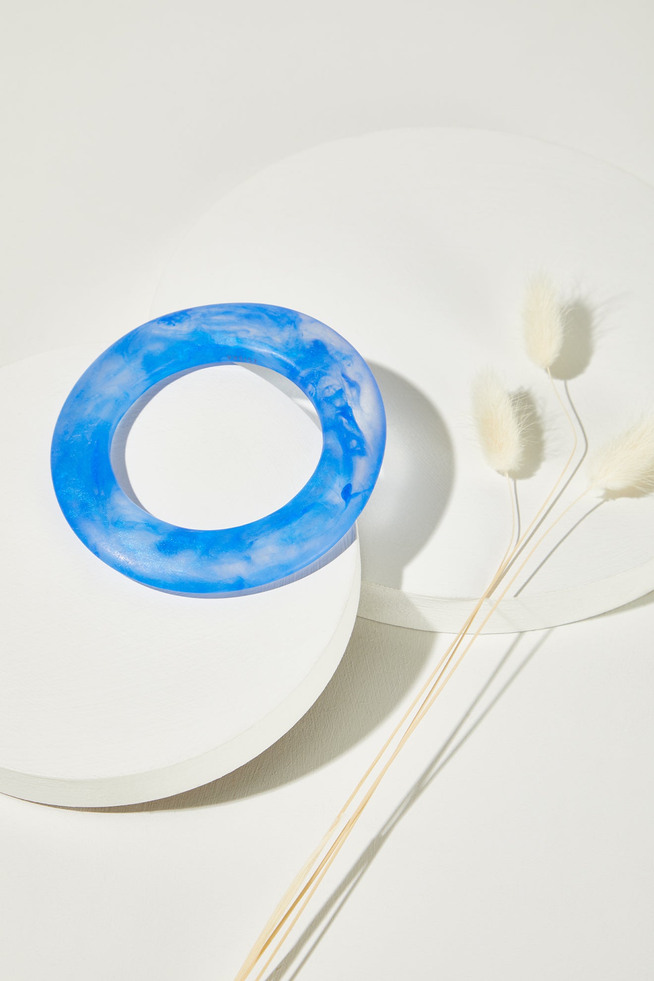 The Marbelled Bracelet, a round blue resin accessory, rests on a white circular platform beside three stems of white dried bunny tail grass.