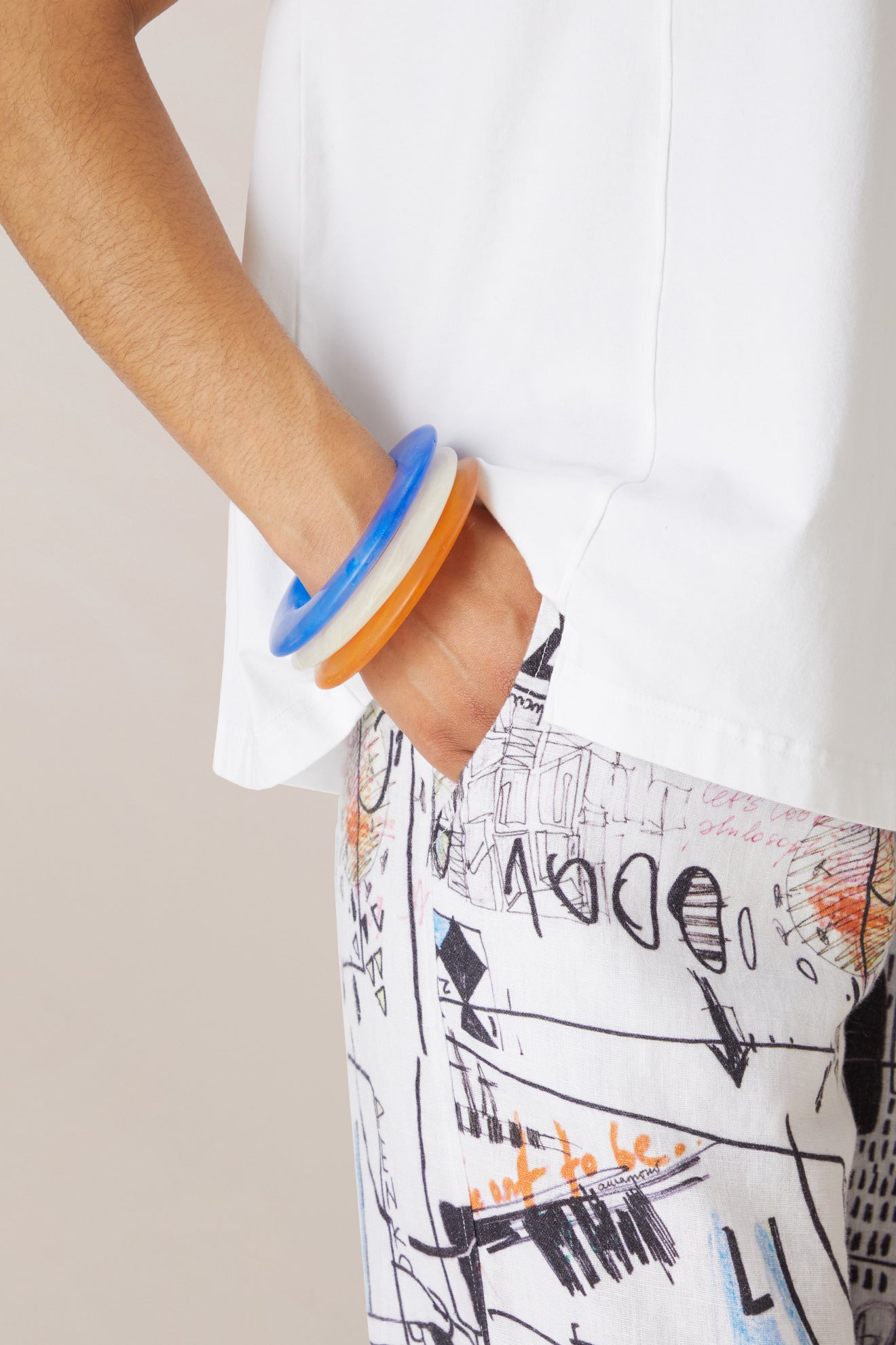 Wearing a white shirt and abstract patterned pants, a person stands with one hand in their pocket, accessorized with two chunky Marbelled Bracelets—one blue and one orange.